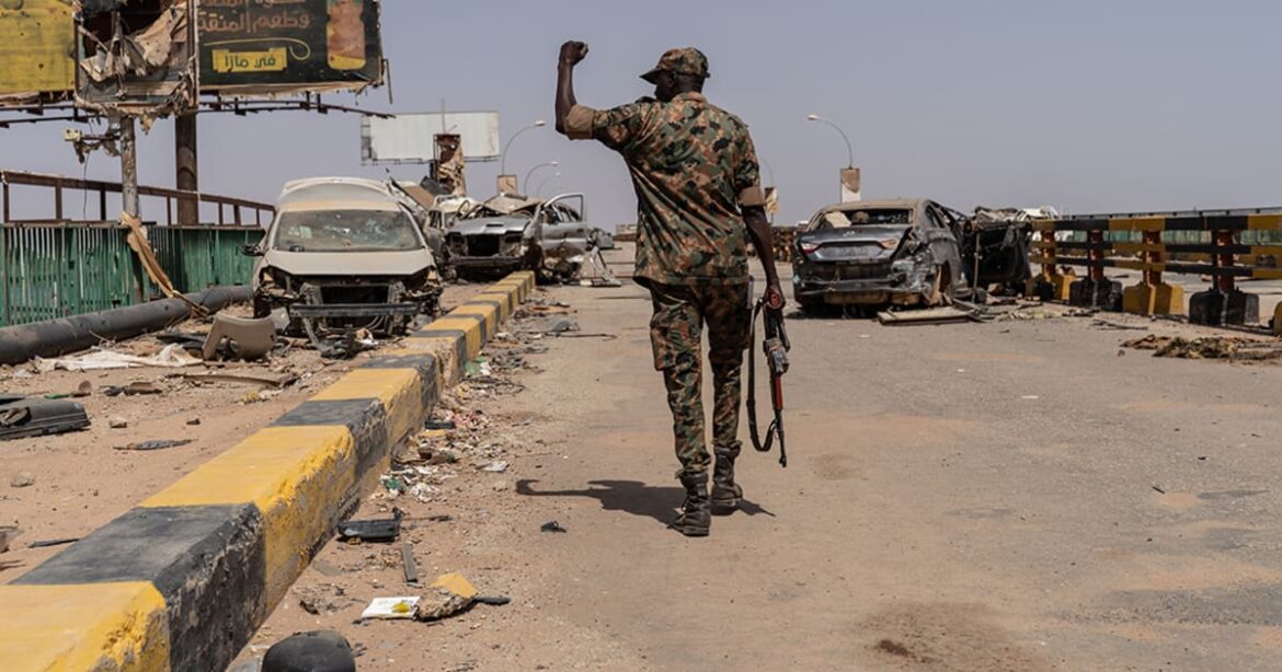 Soldier in camouflage walks along a destroyed highway, raising a clenched fist while holding a rifle; burnt cars and debris litter the scene.