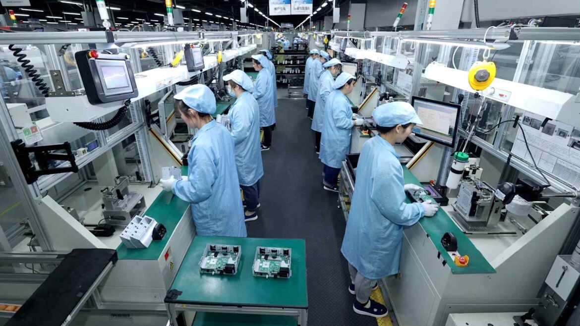 Factory assembly line with workers in blue cleanroom coats assembling electronics.