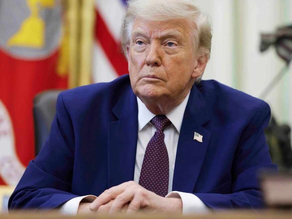 Older man in a navy suit and red patterned tie sits at a desk with his hands folded.