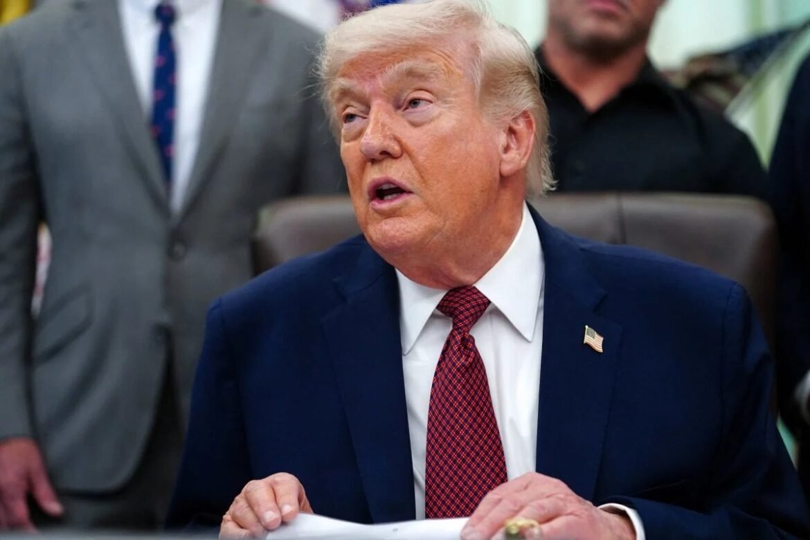 Former US President Donald Trump seated at a desk, wearing a navy suit and red tie with an American flag pin, speaking or signing a document