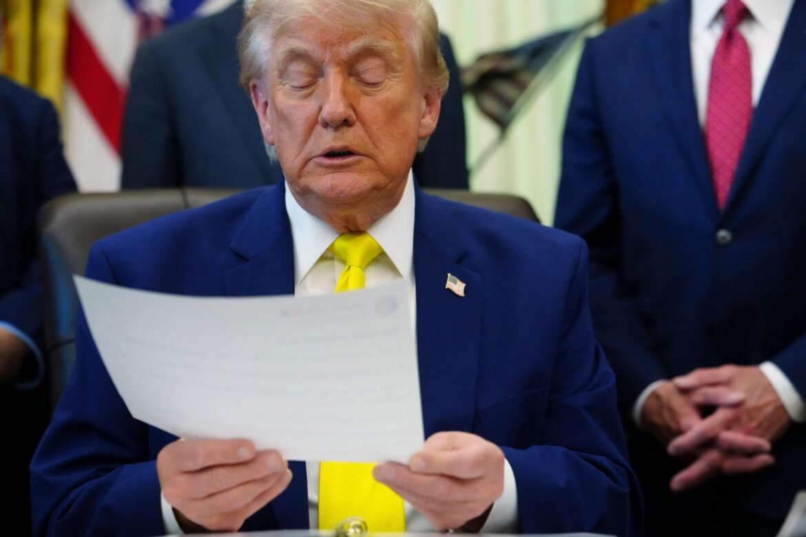 Man in a blue suit with a bright yellow tie reading a document at a formal meeting; American flag lapel pin visible.