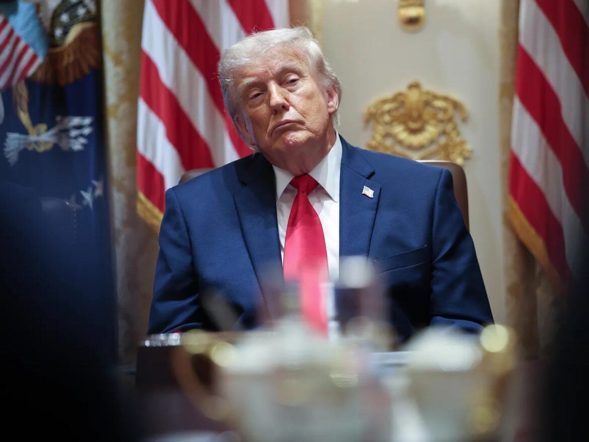 Man in a navy suit and red tie sits at a desk with American flags in the background, looking to the left.