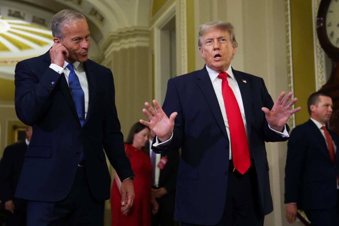 Two men in dark suits and ties stand in a grand hallway; one gestures with his hands while the other adjusts an earpiece.