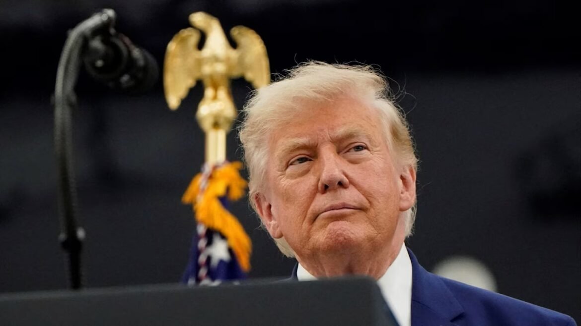 Man in a dark blue suit stands at a podium with a gold eagle emblem behind him, looking upward pensively at a speech event.
