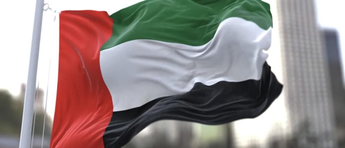 UAE flag waving in the wind on a flagpole, showing a red hoist stripe and horizontal green, white, black stripes.