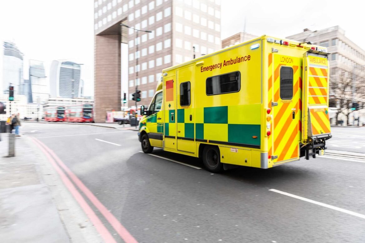 Yellow emergency ambulance driving through a city street with tall office buildings in the background (rear view)