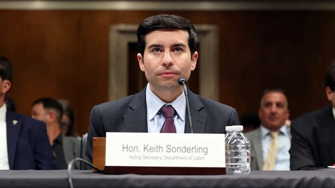 Man in a dark suit testifying at a formal hearing, speaking into a microphone with a water bottle nearby; nameplate reads Acting Secretary, Department of Labor.