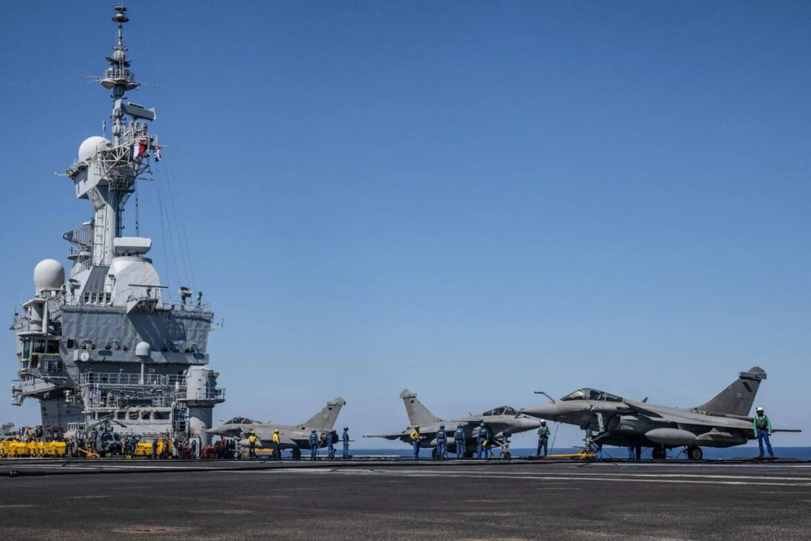 Aircraft carrier flight deck with two gray fighter jets and crew members in blue and yellow vests under a clear blue sky.