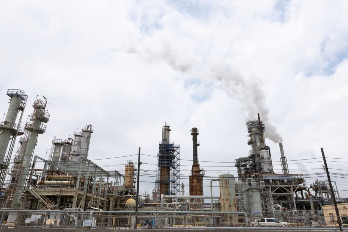 Industrial refinery complex with tall smokestacks emitting white smoke into a cloudy sky.