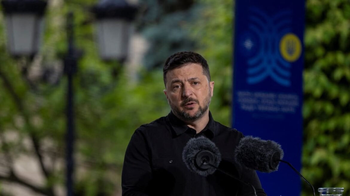 Man in a black shirt speaking at an outdoor press event, two furry microphones in front of him, greenery in the background.