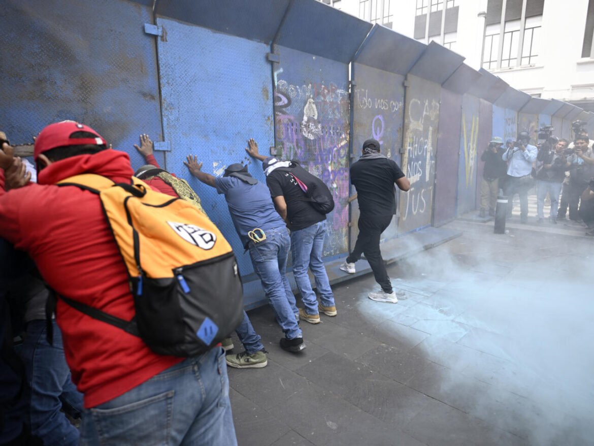 Protesters press against a blue graffiti-covered barrier as smoke billows nearby, with photographers watching.