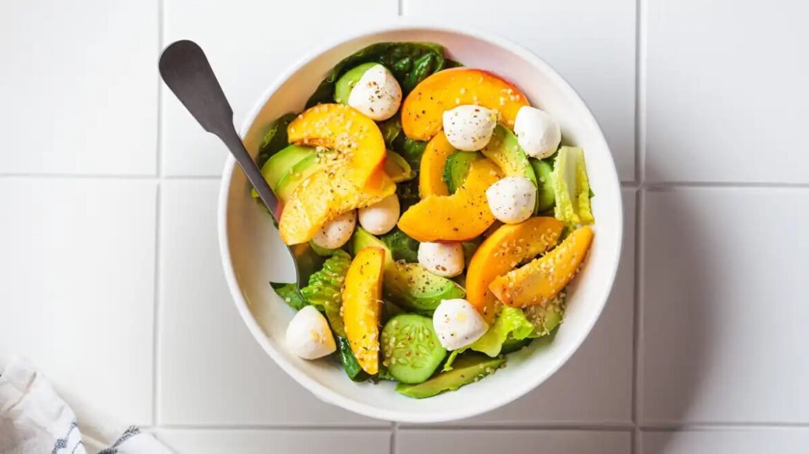 Bowl of mixed salad with peach slices, avocado, cucumber, mozzarella balls, and greens, with a spoon resting in the bowl on a tiled surface.