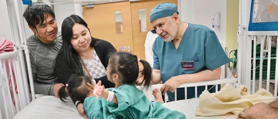 Doctor in teal scrubs talks with a family beside a hospital bed as two young girls in green outfits interact nearby in a pediatrics ward setting.