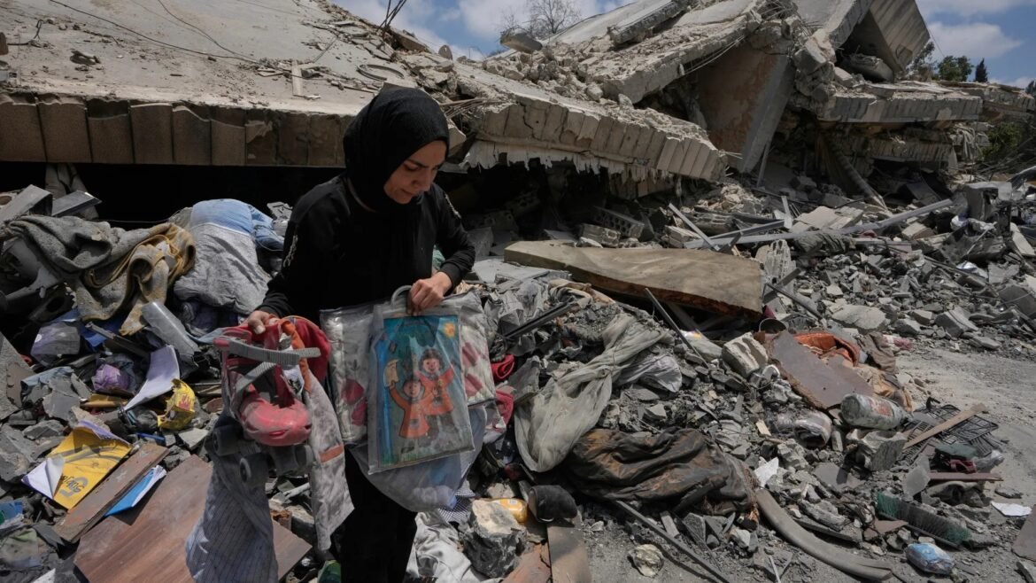 Woman in a black hijab searches through rubble, picking up damaged bags amid destroyed buildings after a disaster.