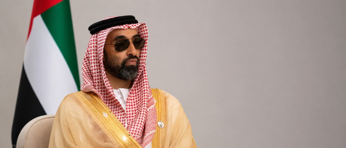 Man in traditional Arab attire (keffiyeh and thobe) wearing sunglasses, seated indoors with a flag in the background.