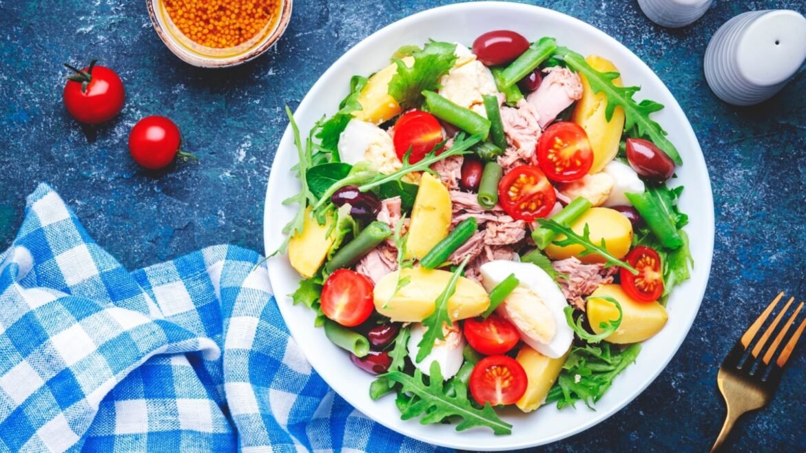 Plate of mixed salad with arugula, cherry tomatoes, potatoes, tuna, olives, green beans and boiled eggs on a white dish and blue tablecloth.