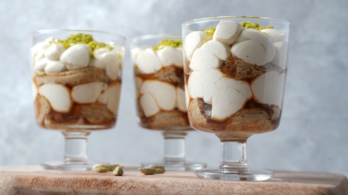 Glass dessert cups on a wooden board, layered with cream, cake, caramel, and crushed pistachios on top.