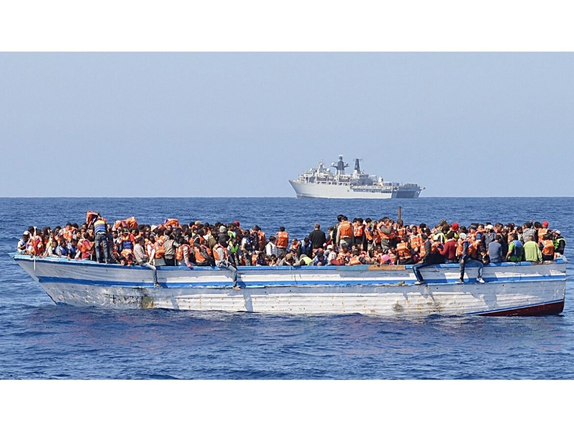 Overcrowded boat at sea filled with passengers wearing orange life jackets; a larger vessel is visible on the horizon in the distance.