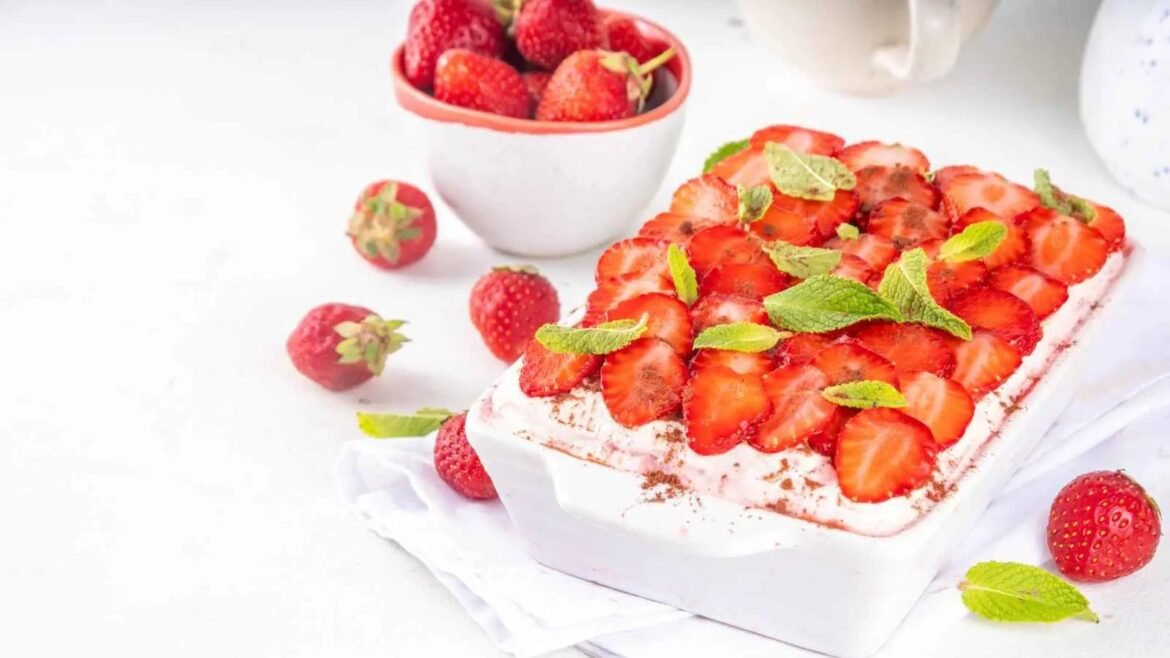 Square dessert topped with strawberry slices, mint leaves, and a cocoa dusting on a white surface.