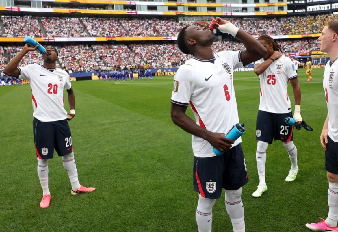 England players drink from water bottles on the field during a match, with a packed stadium in the background.