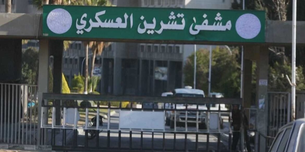 Security checkpoint entrance with a green banner in Arabic above a gate, round white logos on each side, and vehicles in the background.