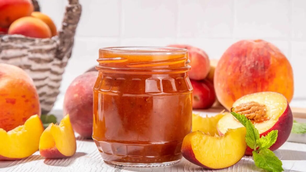 Jar of peach jam on a light wooden table with fresh peach slices and a basket of peaches in the background.