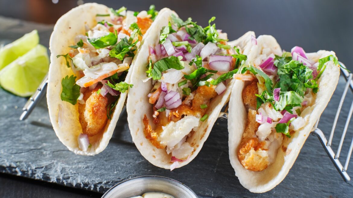Three soft tacos filled with crispy fried fish, shredded vegetables, cilantro, and a creamy sauce, with lime wedges in the background.