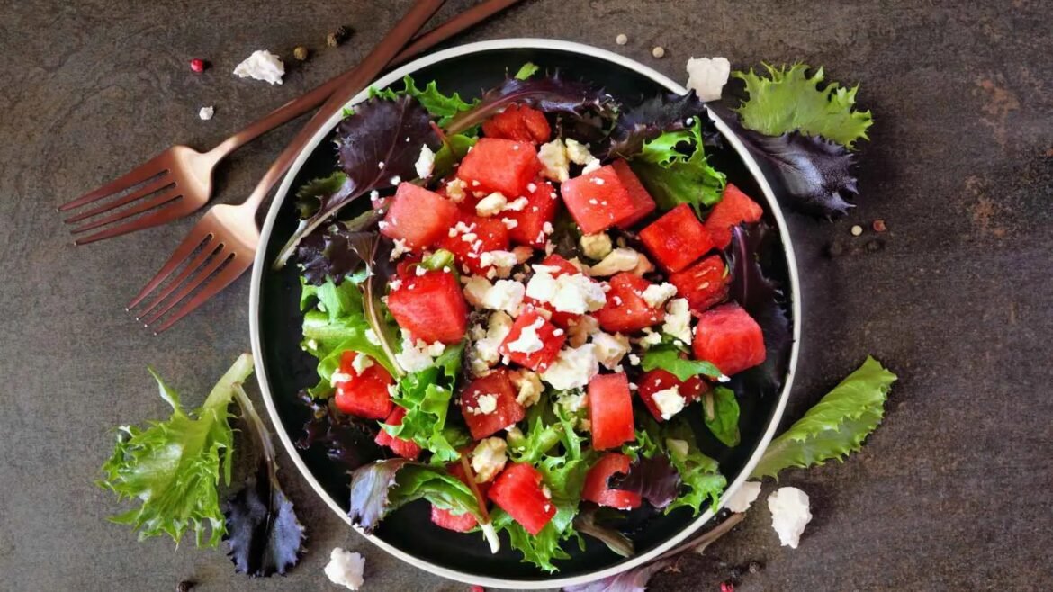 Watermelon and feta salad with mixed greens in a black bowl on a dark surface, crumbled cheese sprinkled on top.
