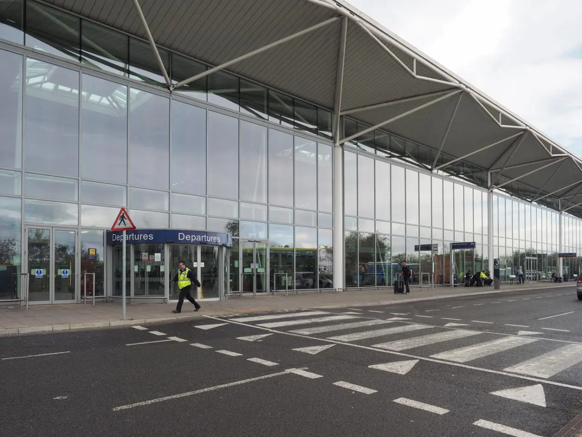Glass-front airport terminal with Departures sign and a pedestrian crossing outside the entrance.