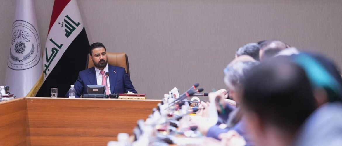 Iraqi official in a navy suit sits at a wooden conference desk, addressing a meeting with attendees on the right.