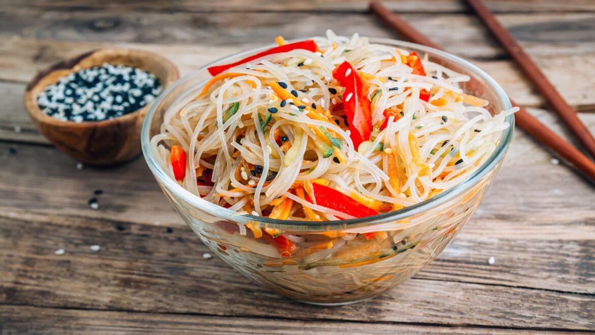 Glass noodle salad with red peppers, carrots, and sesame seeds in a clear bowl on a rustic wooden table.