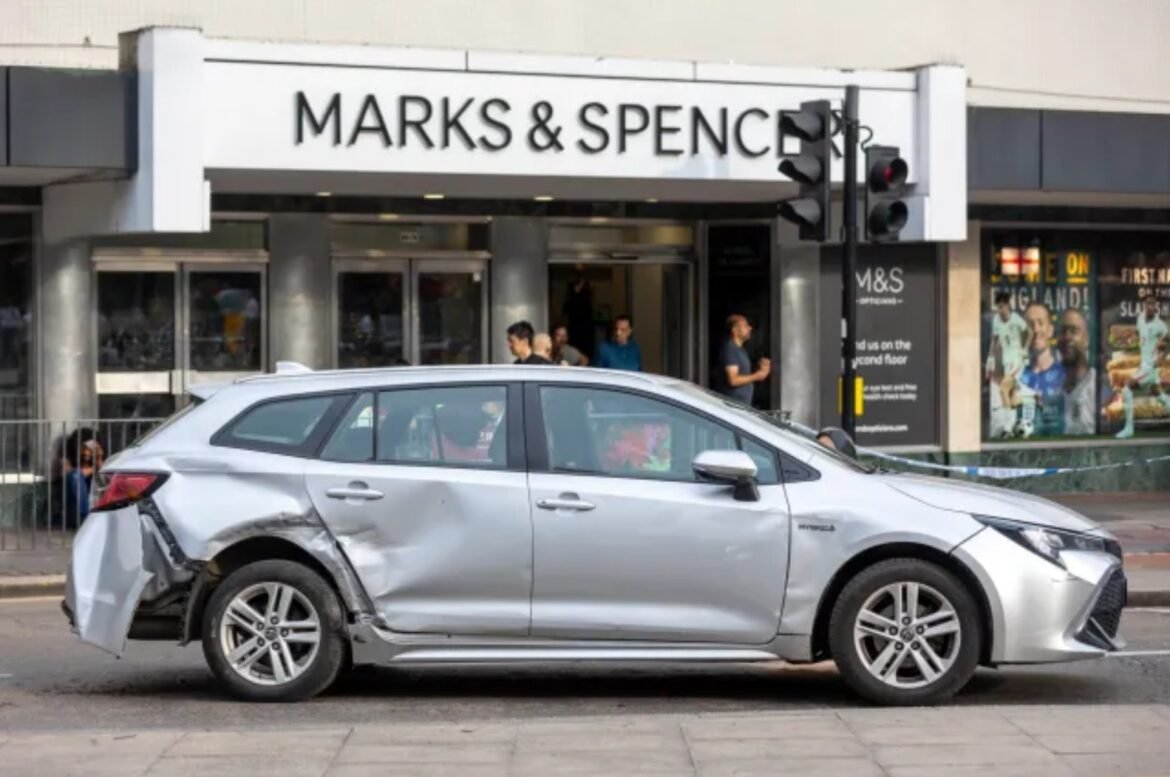 Silver car with extensive side damage parked on a city street in front of a Marks & Spencer store; pedestrians look on in the background.