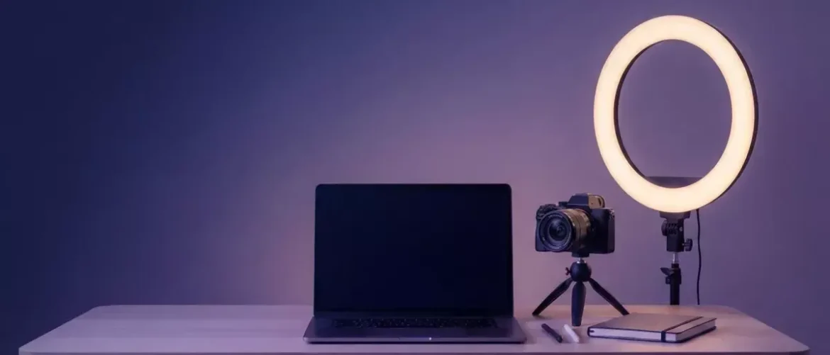 Desk setup with black laptop, DSLR on a small tripod, and a circular ring light beside a notebook and pens on the desk.