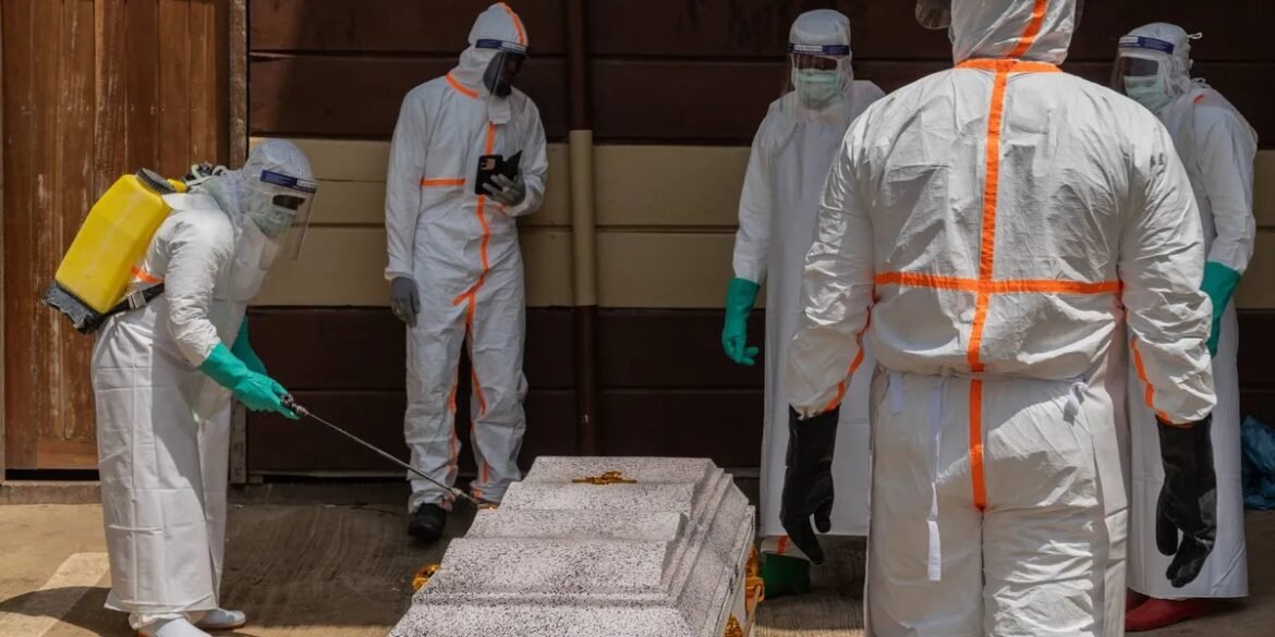 Hazmat-suited team disinfects a casket with a spraying wand in an industrial setting.