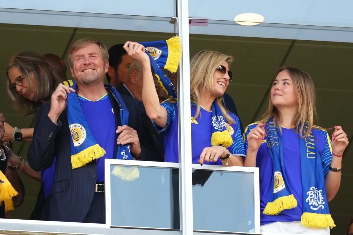 Three fans in blue and yellow attire cheer from a balcony during a soccer match.