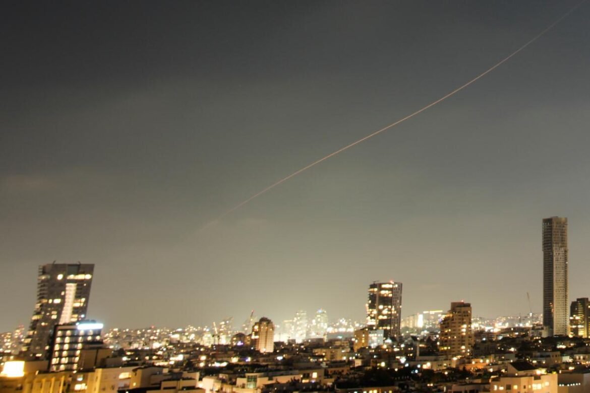 Nighttime city skyline with illuminated buildings and a diagonal contrail across the sky over the urban horizon.