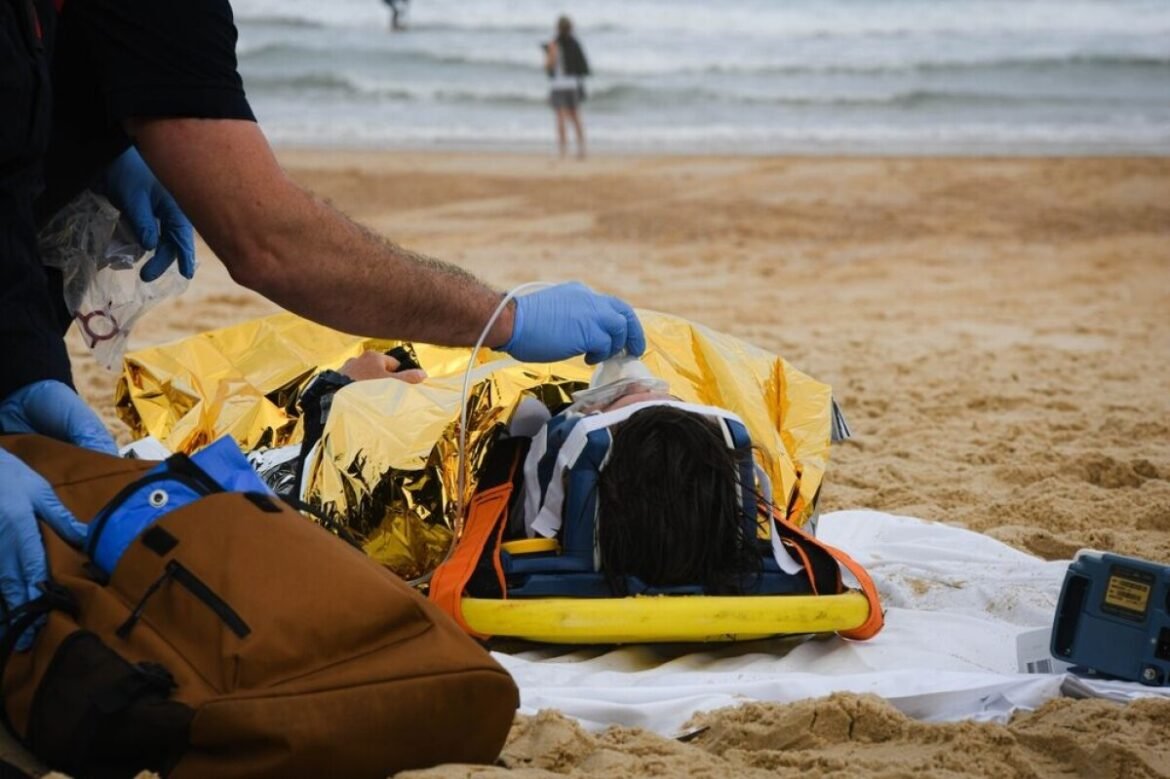 Paramedics in blue gloves attend to a person on a yellow spine board wrapped in a reflective emergency blanket on a sandy beach.
