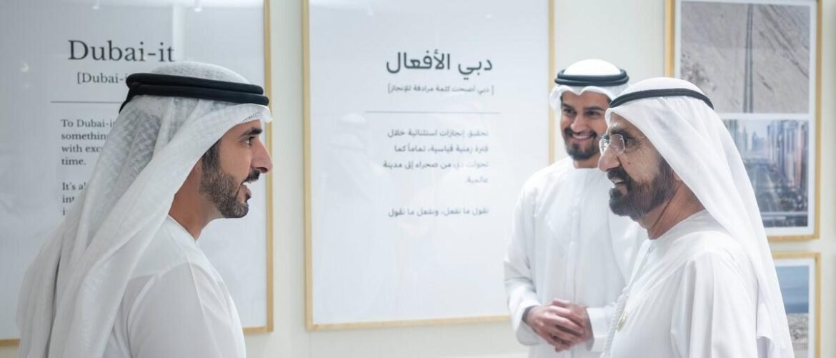 Three men in traditional Emirati white kanduras converse in a bright gallery, with framed Dubai-it posters on the wall behind them.