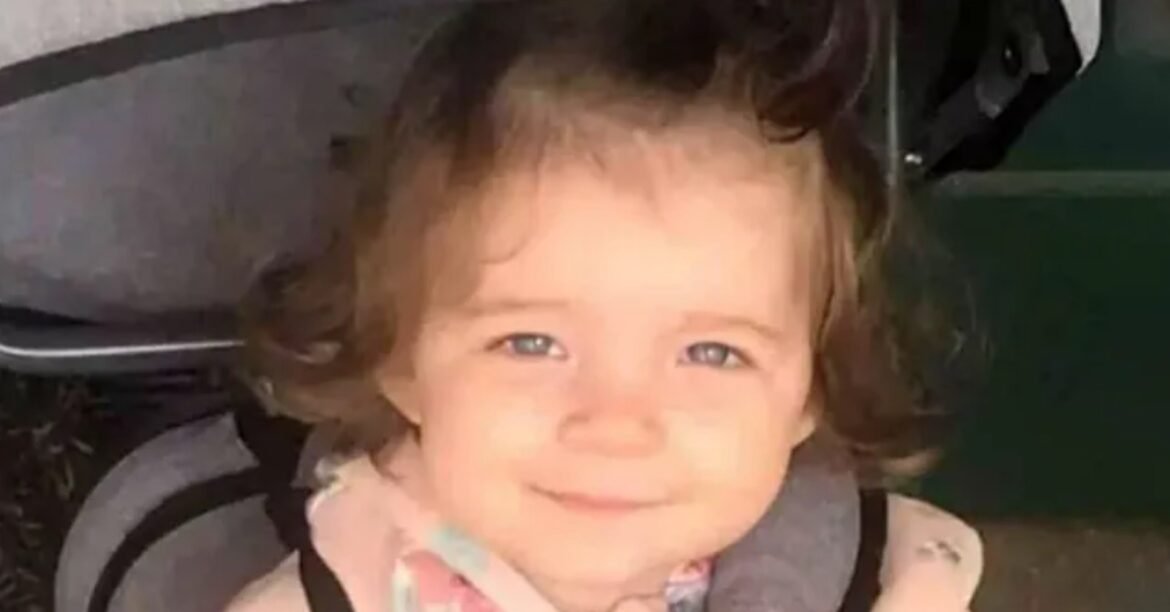 Smiling toddler with brown hair sits in a stroller, looking at the camera wearing a colorful scarf around the neck.