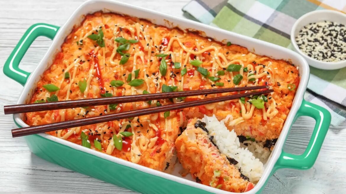 Green ceramic baking dish filled with a spicy salmon bake topped with mayo drizzle, sesame seeds, and chopped green onions; chopsticks rest on top.
