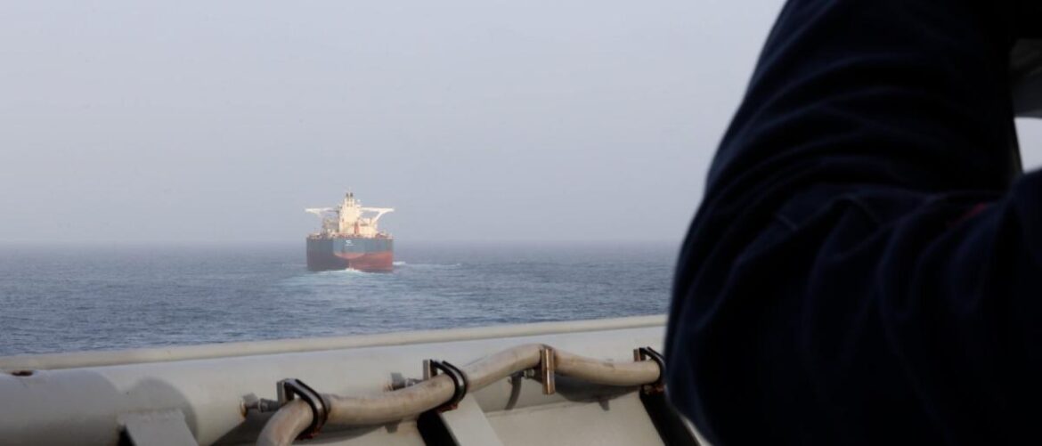 A large cargo ship on calm open water seen from a nearby vessel, with part of a person in dark clothing at the right foreground.