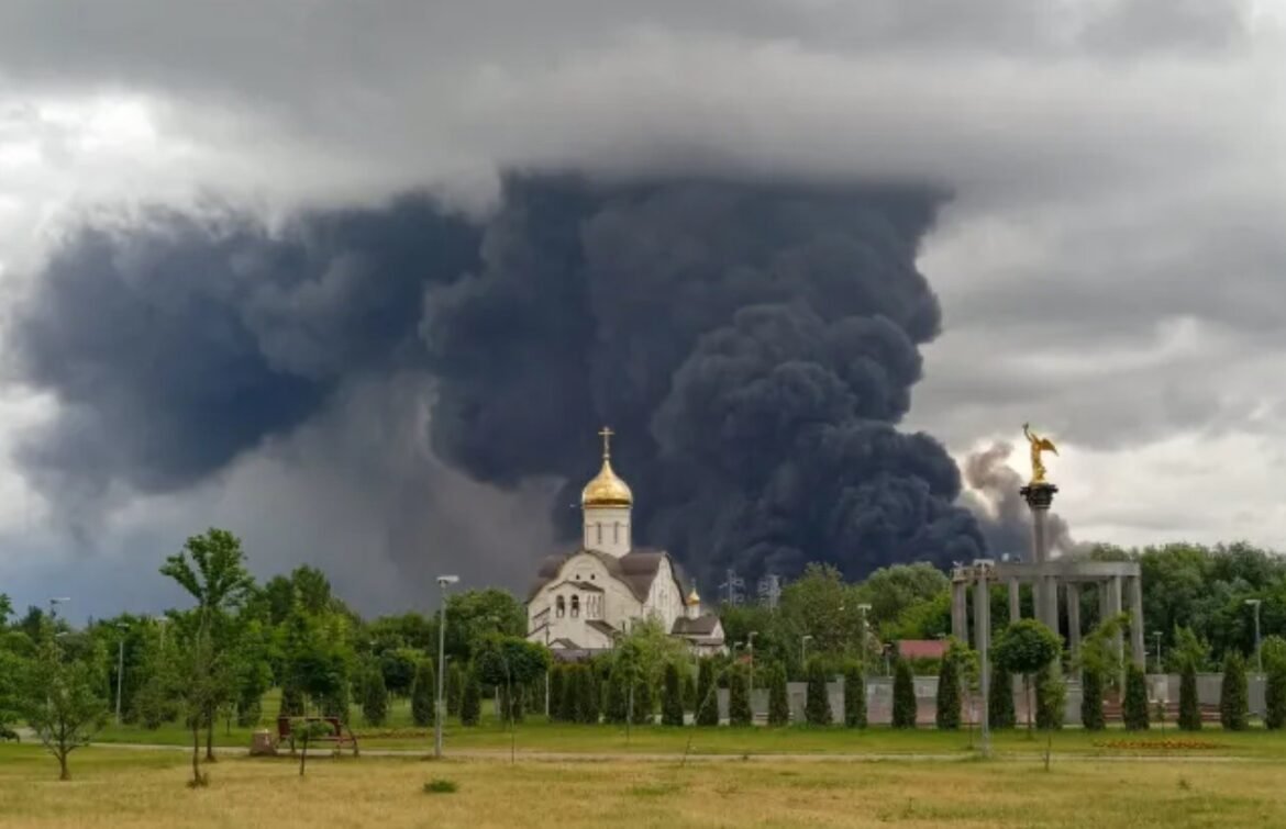 Huge dark smoke plume rises behind a church with a gold dome; a park with trees is in the foreground and a statue stands on a column to the right.