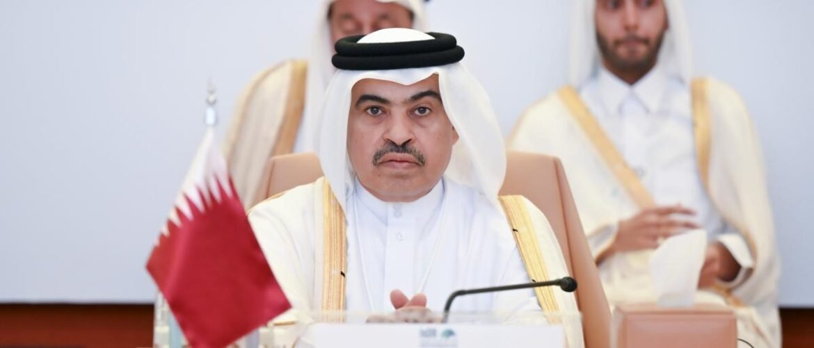 Senior official wearing traditional Qatari attire sits at a conference table with a Qatar flag visible, looking serious during a formal meeting.