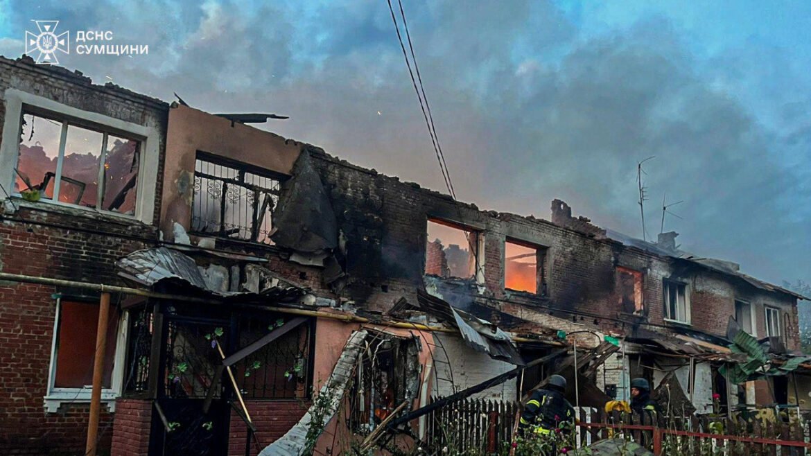 Two-story row house charred by fire, with blown windows and collapsed roof; firefighters in front amid rising smoke. (Emergency services logo in corner)