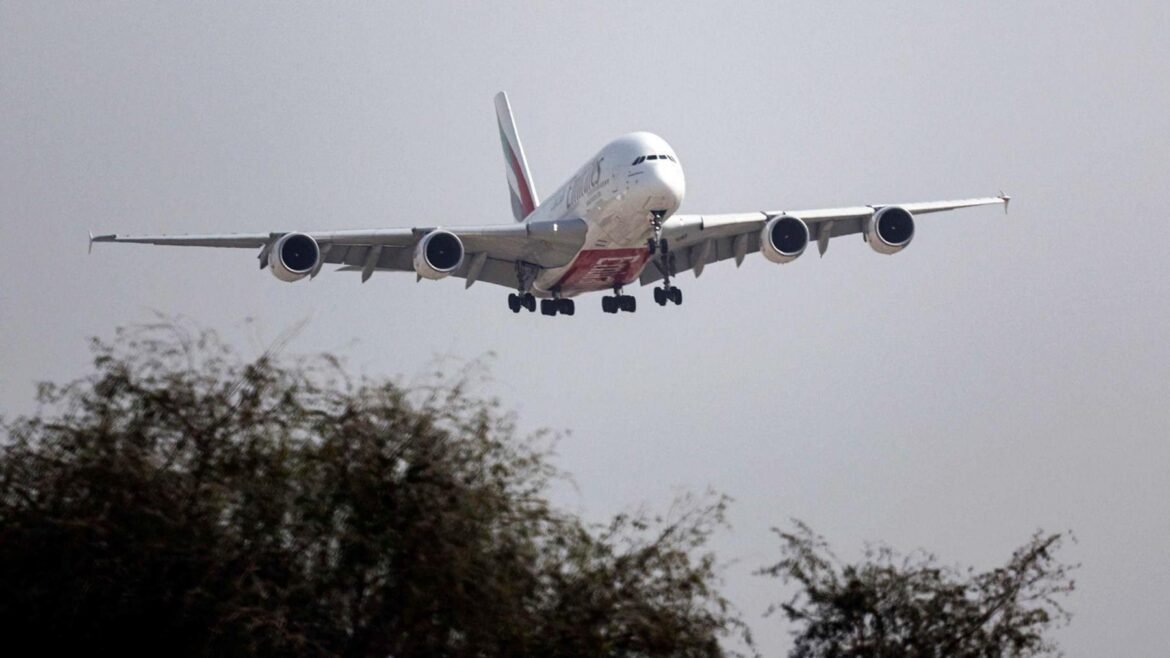 Large four-engine passenger airliner with landing gear down, approaching runway from ground level, trees in foreground