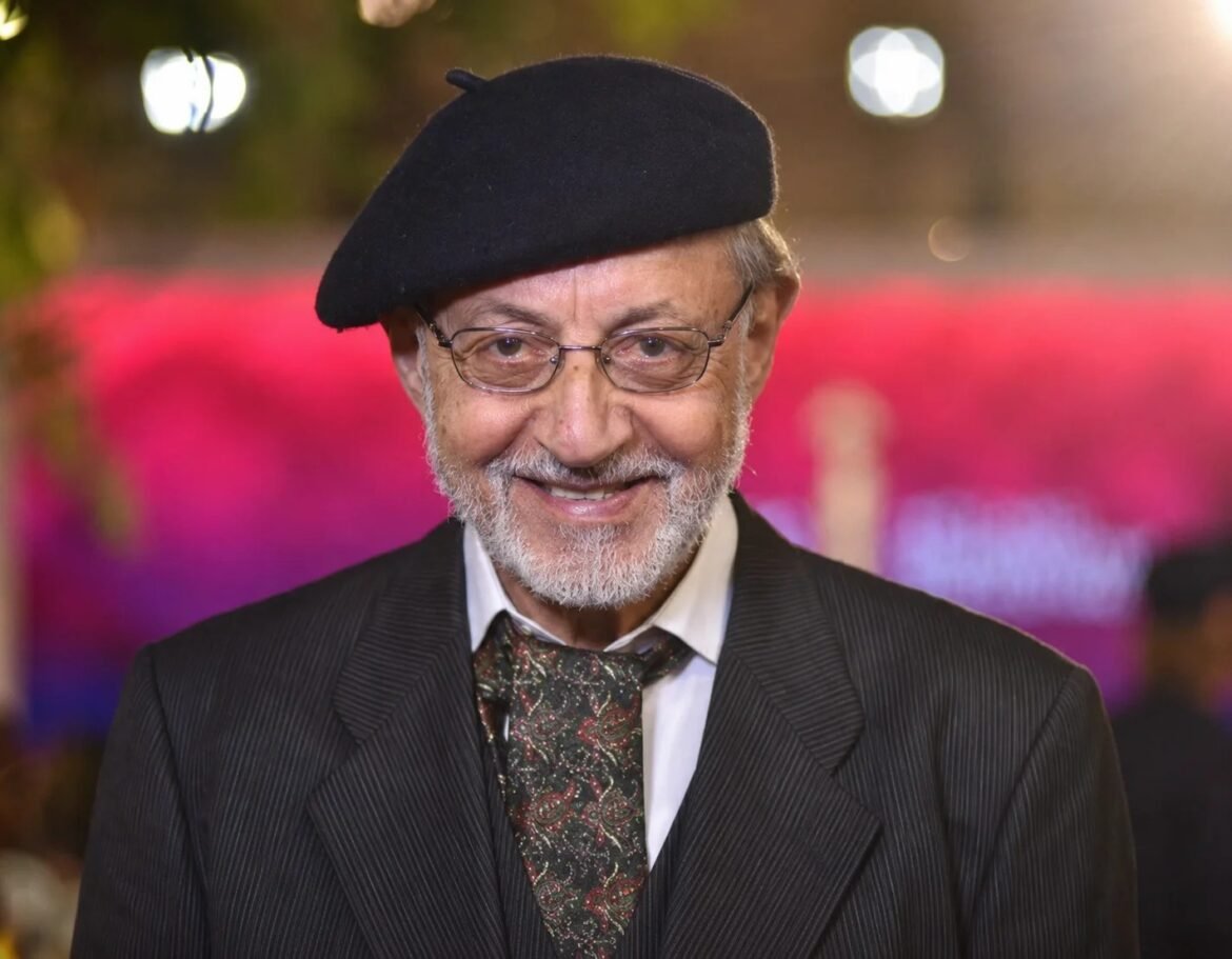 Smiling elderly man with a gray beard wearing a black beret, glasses, and a pinstripe suit at an event.