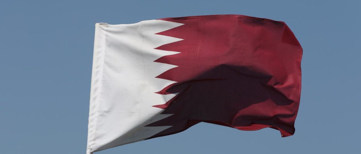 Flag of Qatar fluttering in the wind against a clear blue sky, with a white zigzag border separating the white and maroon fields.