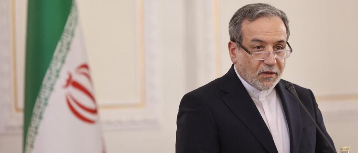 Iranian official in a dark suit and glasses speaks at a podium with the Iranian flag in the background.