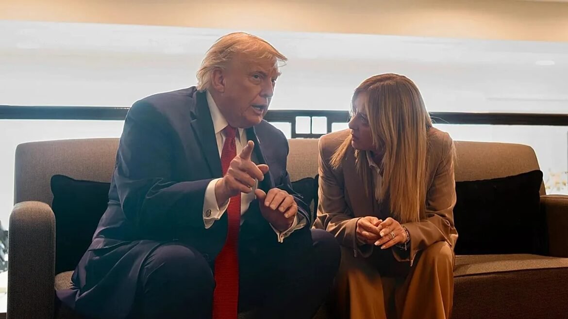 Two people sit on a sofa engaged in a serious conversation; the man in a dark suit and red tie points as he speaks to the woman in a beige suit.