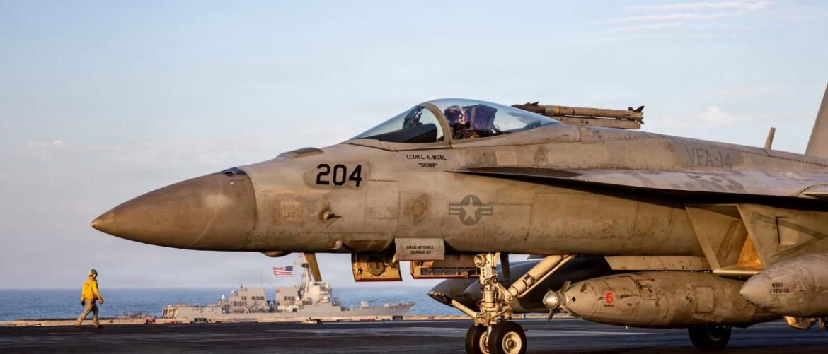 Military jet parked on an aircraft carrier deck with crewman walking in the foreground and a ship visible in the background, under a blue sky